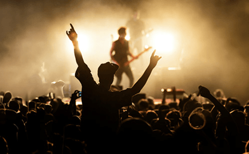 Silhouette of a cheering crowd at a live concert with a band performing on a dimly lit stage.