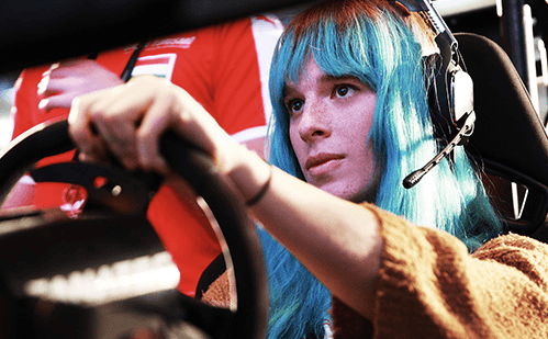 A woman with blue hair plays a car racing game using a steering wheel.