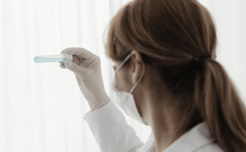 A scientist in a lab coat, mask, and gloves holding up a test tube with blue liquid for inspection.